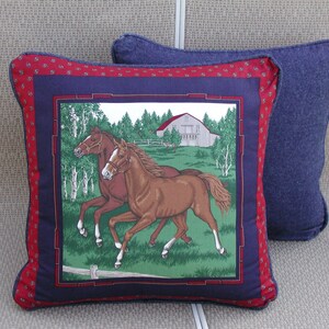 May include: A square decorative pillow featuring a central graphic of two brown horses running in a green field with a white barn in the background. The image is framed by a dark blue border and an outer red border with a small repeating pattern.