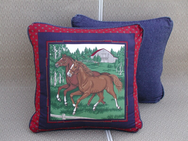 May include: A square decorative pillow featuring a central graphic of two brown horses running in a green field with a white barn in the background. The image is framed by a dark blue border and an outer red border with a small repeating pattern.