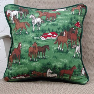 May include: A square throw pillow featuring a green fabric cover with a repeating pattern of brown and white horses in a pasture. The design includes small red-roofed farm buildings and green trees. The pillow is finished with dark green piping.