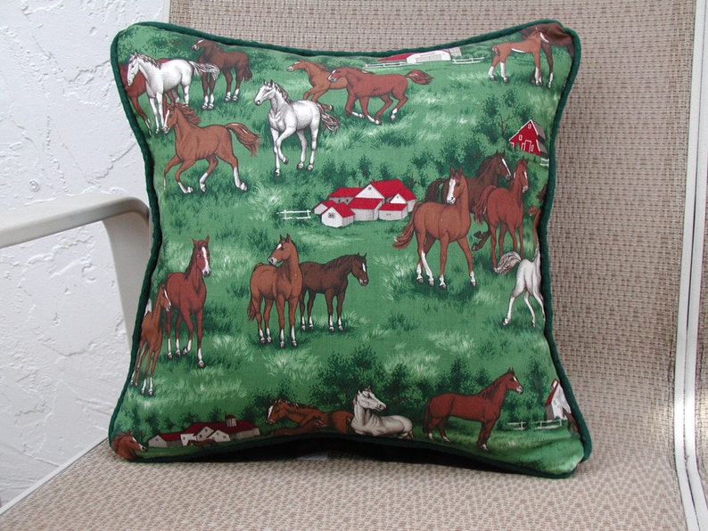 May include: A square throw pillow featuring a green fabric cover with a repeating pattern of brown and white horses in a pasture. The design includes small red-roofed farm buildings and green trees. The pillow is finished with dark green piping.