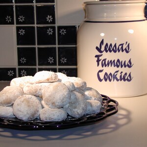 Op de afbeelding: Een witte keramische koekjespot met blauwe letters die "Leesa's Famous Cookies" lezen. Een bord met witte, met poedersuiker bestoven koekjes staat op een zwart aanrecht.