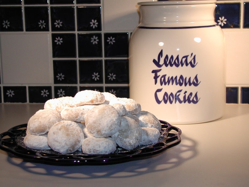 Op de afbeelding: Een witte keramische koekjespot met blauwe letters die "Leesa's Famous Cookies" lezen. Een bord met witte, met poedersuiker bestoven koekjes staat op een zwart aanrecht.