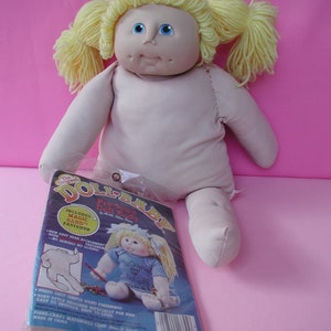 Puede incluir: Una muñeca rosa y blanca con cabello rubio y ojos azules. La muñeca lleva un vestido rosa y está sentada sobre una superficie rosa. La muñeca sostiene un paquete blanco con las palabras "The Original Doll Baby Pre-Sewn Doll Body" impresas en él.