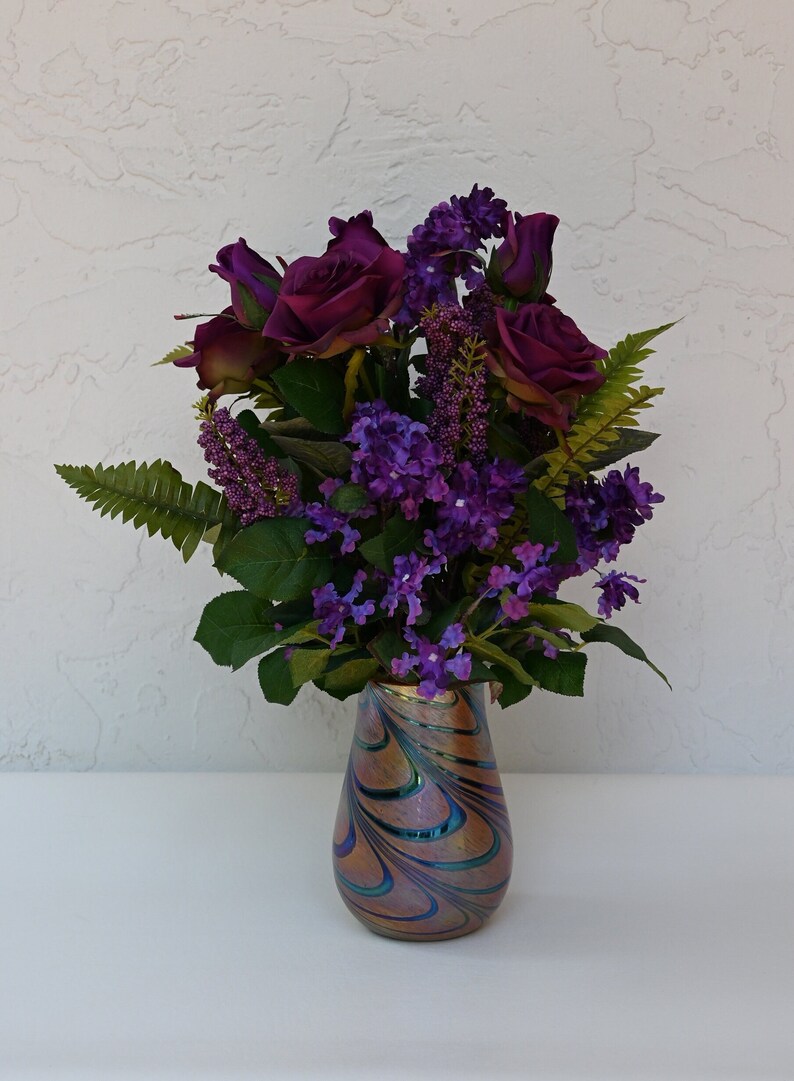 May include: A bouquet of purple roses and lilac flowers in a iridescent glass vase with swirling patterns.