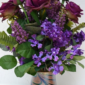 May include: A bouquet of purple roses and lilac flowers in a swirled glass vase with a brown, blue, and green pattern.