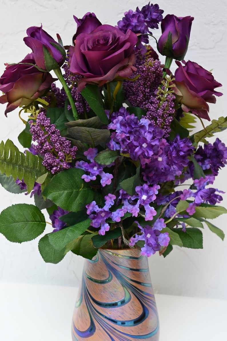 May include: A bouquet of purple roses and lilac flowers in a swirled glass vase with a brown, blue, and green pattern.