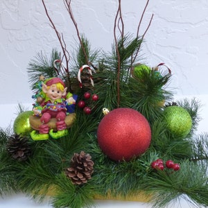 Puede incluir: Centro de mesa navideño festivo con una figura de elfo sonriente, adornos rojos y verdes, piñas y ramitas de bayas rojas. El arreglo incluye bastones de caramelo y está sobre ramas de hoja perenne.