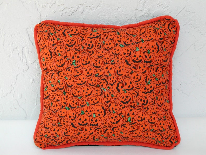K&ouml;nnte beinhalten: Ein orangefarbenes Kissen mit Halloween-Motiv, das ein sich wiederholendes Muster aus schwarzen K&uuml;rbisgesichtern auf schwarzem Hintergrund zeigt. Das Kissen hat einen roten Rand.