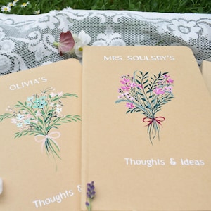 May include: Two beige notebooks with floral illustrations and text. One notebook reads "OLIVIA'S" with a bouquet of blue, pink, and green flowers tied with a pink ribbon. The other reads "MRS SOULSBY'S" with a bouquet of pink flowers tied with a red ribbon. Both read "Thoughts & Ideas".