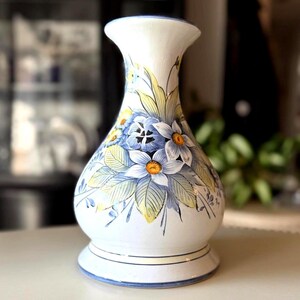 May include: A white ceramic vase with a bulbous base and flared top. The vase is decorated with blue and white flowers, yellow accents, and green leaves. The vase has a blue rim and base trim.