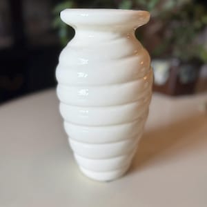 May include: A white ceramic vase with a textured, ridged design. The vase has a wide base that tapers to a narrow neck and flared rim. The vase is sitting on a white surface.