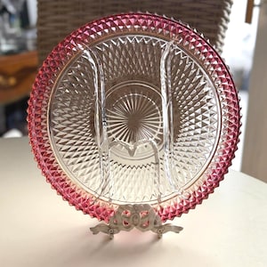 May include: A clear glass serving dish with a pink rim and diamond pattern. The dish is divided into sections and sits on a decorative metal stand. The rim has a textured, pointed design.