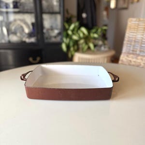 May include: A square, brown and white enamel baking dish with handles. The dish has a white interior and a brown exterior. The handles are also brown. The dish is sitting on a white table.
