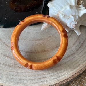 May include: An orange-brown bangle bracelet with carved floral designs. The bracelet is made of a glossy material and is displayed on a wooden surface. A seashell and ceramic piece are in the background.