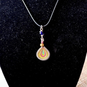 May include: A handcrafted pendant necklace featuring a teardrop-shaped pendant with a colorful, intricate design. The pendant is suspended from a gray cord and includes blue and brown beads. The pendant's design incorporates yellow, pink, and green hues.