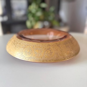 May include: A decorative, low, round dish with a gold-colored, ornate, floral pattern around the rim. The center of the dish is a translucent, amber color. The dish is likely made of glass or crystal.