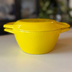 May include: A bright yellow, enameled cast iron Dutch oven with a matching lid. The pot has two side handles and a rounded shape. The lid has a handle and fits snugly on top. The pot is sitting on a white surface.