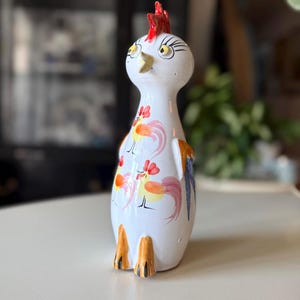 May include: A white ceramic rooster figurine with a red comb, yellow beak, and orange feet. The rooster has painted details including floral designs in red, orange, and yellow. The figurine is approximately 8 inches tall.