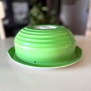 May include: A bright green ceramic cloche with a matching base. The cloche has a rounded, ridged design and a small hole at the top. The base is white with a green rim. The cloche is sitting on a white surface.