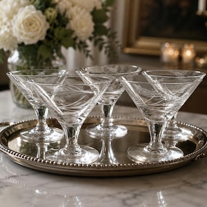 May include: A set of four clear glass martini glasses with etched floral patterns on the bowls, each featuring a short stem and a circular base, arranged on a round silver-toned metal serving tray with a beaded edge and two side handles.
