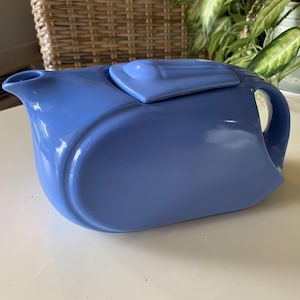 May include: A light blue ceramic teapot with a matching lid. The teapot has a curved, streamlined design with a handle and spout. The teapot is sitting on a white surface.