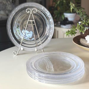 May include: A set of clear glass plates featuring a textured beaded rim pattern. One plate is displayed upright on a silver metal stand, while three additional plates are stacked horizontally in front of it, showcasing the consistent circular design.