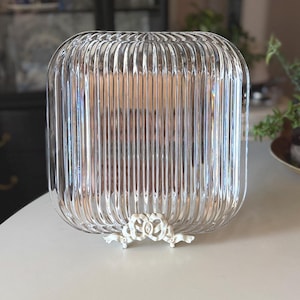 May include: A square, clear glass plate featuring a vertical ribbed texture throughout its surface. The plate is supported at the bottom by a decorative, off-white metal stand that displays an ornate, bow-like design with intricate detailing.