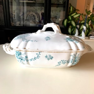 May include: White porcelain serving dish with a lid, featuring a teal floral pattern and gold trim. The dish has a handle on the lid and handles on the sides. A classic piece for serving food.