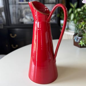 May include: A vibrant red ceramic pitcher with a curved handle and a slightly flared spout. The pitcher has a glossy finish and a gold-colored rim. It is a decorative item, likely for serving beverages or displaying flowers.