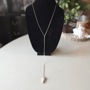 May include: A silver-toned necklace with a Y-shaped design, displayed on a black mannequin. The necklace features a delicate chain that culminates in a teardrop-shaped pendant with a white stone. The necklace is presented on a white surface.