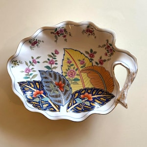 May include: A white ceramic leaf-shaped serving dish with a scalloped edge and a handle. The dish features a floral and leaf design in yellow, orange, blue, and pink, with gold accents. The rim is outlined in blue.
