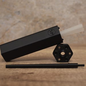 May include: A black rectangular object with a white tube extending from it, a black hexagonal piece, and a black rod. The rectangular object has the letters "SP" on it. The items are on a wooden surface.