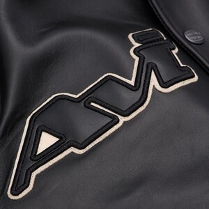 May include: Close-up of a black leather jacket with a large, embroidered patch. The patch features a stylized design in black with a cream-colored border. A button is visible on the upper right side of the jacket.