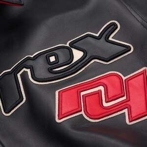 May include: Close-up of a black leather jacket with embroidered lettering. The letters "RS" are in red with a cream border, and the letters "DX" are in black with a cream border. The jacket has a red collar.