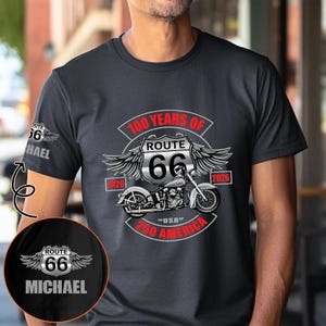 Puede incluir: Camiseta gris oscuro con un diseño de motocicleta de la Ruta 66. El diseño incluye el texto "100 Years of Route 66" y "250 America". La manga tiene un logotipo de la Ruta 66 con el nombre "MICHAEL".