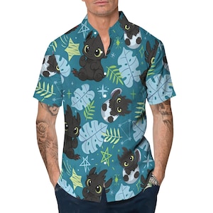 May include: Teal button-up shirt featuring a repeating pattern of black cartoon dragons, white eggs, green leaves, and yellow stars. The short-sleeved shirt has a classic collar and is made of a lightweight fabric.