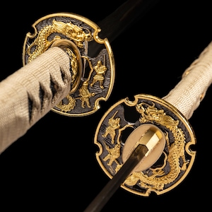May include: Close-up of a Japanese katana sword. The sword features a gold dragon and warrior design on the handguard. The handle is wrapped in a cream fabric. The blade is black.