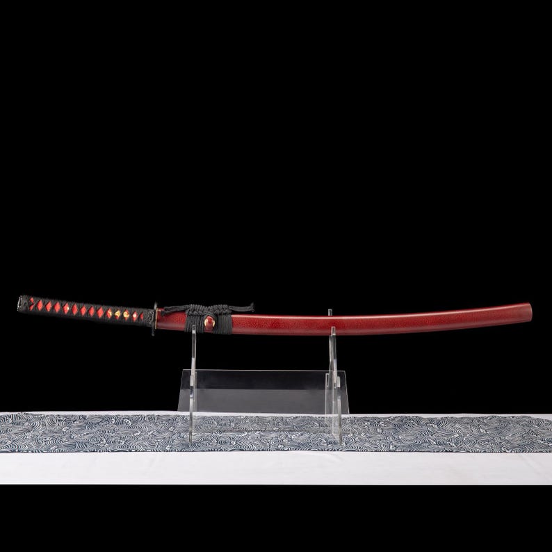 Peut inclure: Un katana japonais avec un fourreau et un manche laqu&eacute;s rouges. Le manche est envelopp&eacute; d'un cordon noir avec des accents rouges en forme de losange. L'&eacute;p&eacute;e est expos&eacute;e sur un pr&eacute;sentoir en acrylique transparent sur fond noir.