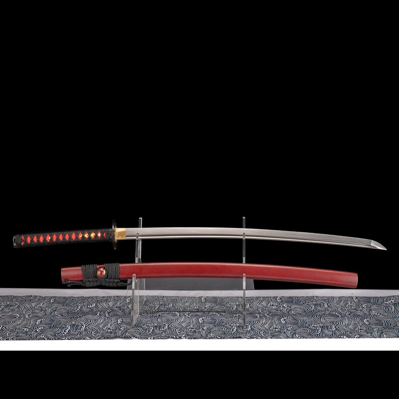 Peut inclure: Un katana japonais avec un manche rouge et noir et une lame argent&eacute;e, expos&eacute; sur un support transparent. Le fourreau rouge est en dessous de la lame. Le fond est noir.