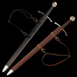 Op de afbeelding: Twee middeleeuwse zwaarden met scheden. Eén zwaard heeft een zwarte leren schede en handvat, de andere heeft een bruine leren schede en handvat. Beide zwaarden hebben zilveren pareerstangen en knoppen, en leren riemen.