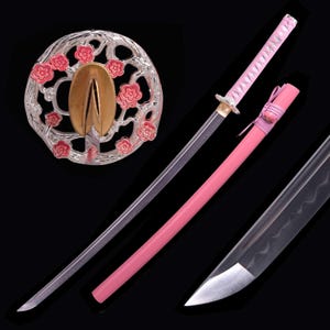 Op de afbeelding: Een roze katana-zwaard met een zilveren lemmet en een roze handvat. De schede is ook roze. De pareerstang is zilver met roze bloemaccenten en een gouden centrum. Het zwaard wordt gepresenteerd tegen een zwarte achtergrond.