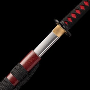 Peut inclure: Gros plan sur un katana. La poign&eacute;e noire est orn&eacute;e d'un motif losange rouge. Le fourreau rouge a une finition textur&eacute;e et la lame est argent&eacute;e. La garde est noire et dor&eacute;e, sur fond noir.