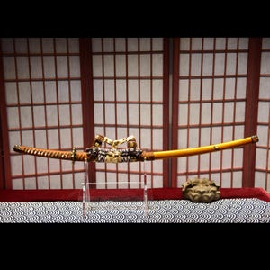 May include: A traditional Japanese sword, or katana, with a brown leather handle and a gold-colored blade. The sword is displayed on a clear stand against a backdrop of a wooden screen.