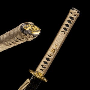 May include: Close-up of a Japanese katana sword. The handle is wrapped in a tan-coloured fabric with black diamond-shaped accents. The guard and pommel feature gold-coloured decorative elements. The blade is black, and the overall design is ornate.