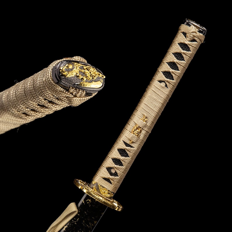 May include: Close-up of a Japanese katana sword. The handle is wrapped in a tan-coloured fabric with black diamond-shaped accents. The guard and pommel feature gold-coloured decorative elements. The blade is black, and the overall design is ornate.