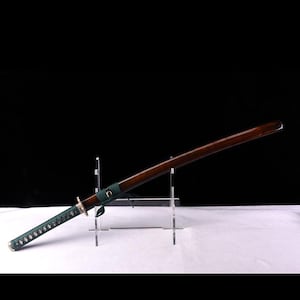 May include: A brown wooden katana sword with a green wrapped handle and a silver metal guard. The sword is displayed on a clear acrylic stand.