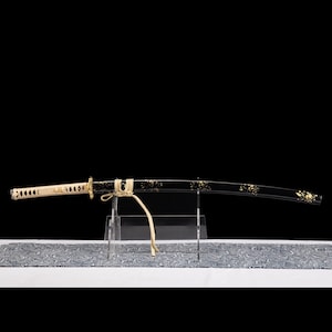 May include: A Japanese katana sword with a black blade and gold floral detailing. The handle is cream-coloured with a diamond pattern. The sword is displayed on a clear stand, with patterned fabric beneath.