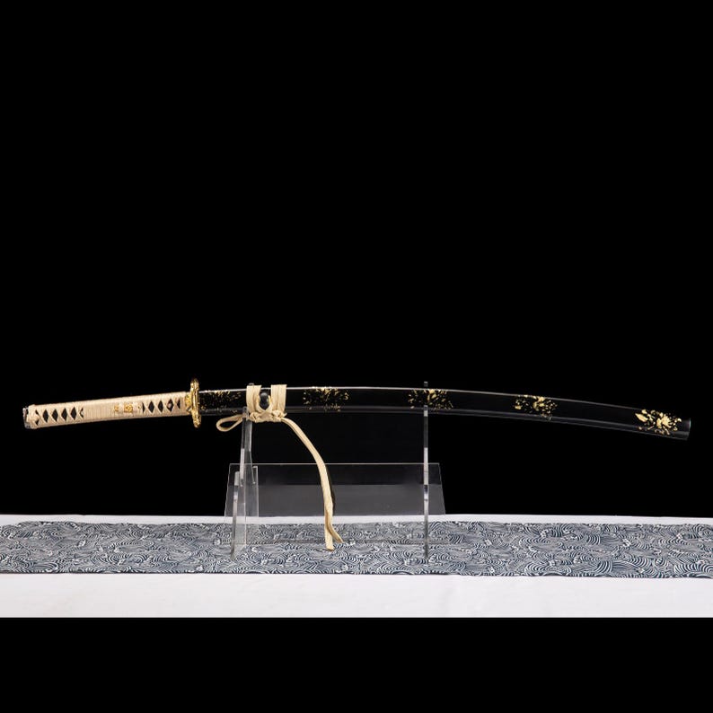 May include: A Japanese katana sword with a black blade and gold floral detailing. The handle is cream-coloured with a diamond pattern. The sword is displayed on a clear stand, with patterned fabric beneath.