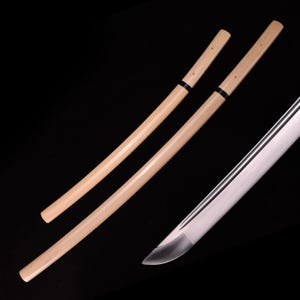 Pode incluir: Duas bainhas de madeira para uma katana. As bainhas são de cor bege claro com detalhes pretos. As bainhas estão sobre uma superfície preta.