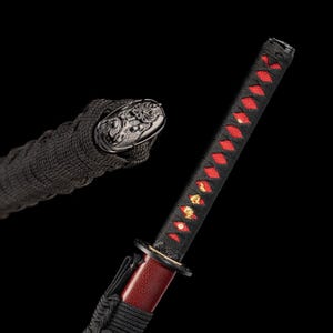 Peut inclure: Gros plan sur un katana japonais. Le manche est envelopp&eacute; de tissu noir avec des losanges rouges. La garde est noire et la lame a une finition rouge. Le pommeau pr&eacute;sente un motif de dragon d&eacute;taill&eacute;.